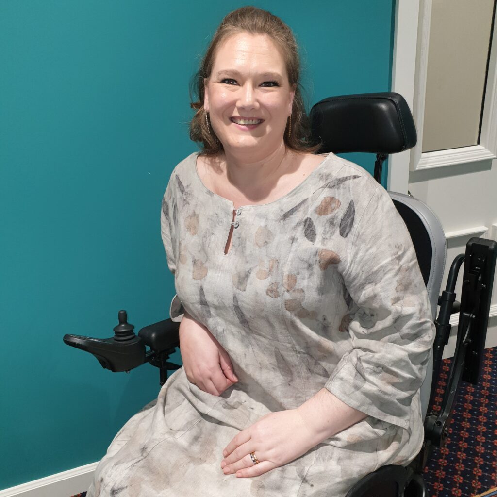 a woman sat in a wheelchair, smiling, wearing a green dress, with leaf shaped patterns on it