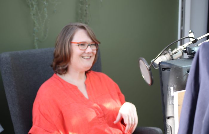 A woman sat in a grey chair, with an orange top on, speaking into a microphone on a video call